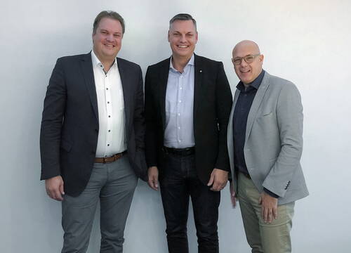 Drei lächelnde Männer in Businesskleidung stehen nebeneinander vor einer hellen Wand und posieren für die Kamera.