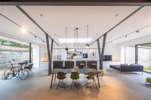 Großer, lichtdurchfluteter Loft-Wohnraum mit offener Küche, langem Esstisch mit bunten Designstühlen, moderner Sofalandschaft, Industriebalken an der Decke und raumhohen Fenstern zum Innenhof.
