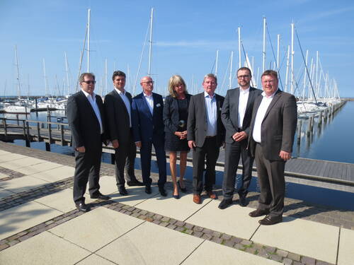 Gruppe von Geschäftsleuten in Anzügen posiert bei sonnigem Wetter auf einer Uferpromenade vor einem Yachthafen mit zahlreichen Segelbooten