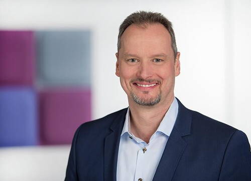 Mittelalterlicher Mann im blauen Business-Anzug lächelt freundlich vor einem unscharfen Hintergrund mit farbigen, quadratischen Wandpaneelen.