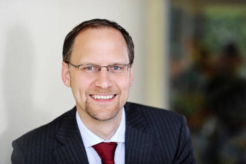 Lächelnder Mann mit Brille im dunklen Nadelstreifenanzug und roter Krawatte vor unscharfem Hintergrund
