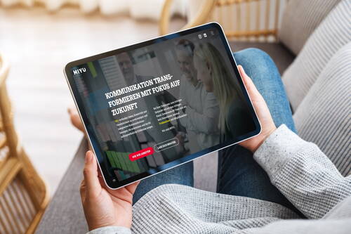 Person sitzt entspannt auf dem Sofa und hält ein Tablet, auf dem eine Website zum Thema zukunftsorientierte Kommunikation mit einem lächelnden Geschäftsteam angezeigt wird.