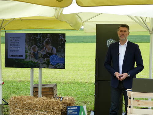 Mann in Anzug präsentiert im Freien neben einem Kühlschrank und einem Bildschirm mit der Aufschrift „Willkommen zur Frische-Expedition“ bei einem Liebherr-Fachpresse-Event auf einem Feld.