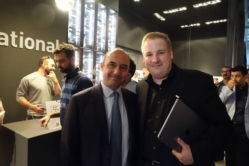 Zwei Männer in Anzug und Hemd lächeln in einer modernen, gut beleuchteten Ausstellungs- oder Messehalle in die Kamera, während im Hintergrund mehrere Besucher miteinander sprechen und ein Mann ein Tablet unter dem Arm hält.