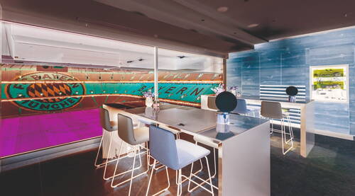 Exklusive Stadionlounge mit modernen Bartischen und Designlampen, die durch eine Glasfront direkten Blick auf die leeren Ränge und das Spielfeld der Allianz Arena mit großem FC‑Bayern-Logo bietet.