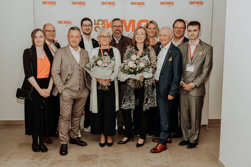 Gruppe festlich gekleideter Mitarbeiter und Gäste posiert lächelnd bei einer Jubiläumsfeier, zwei Frauen in der Mitte halten große Blumensträuße vor einer KMG-Fotowand.