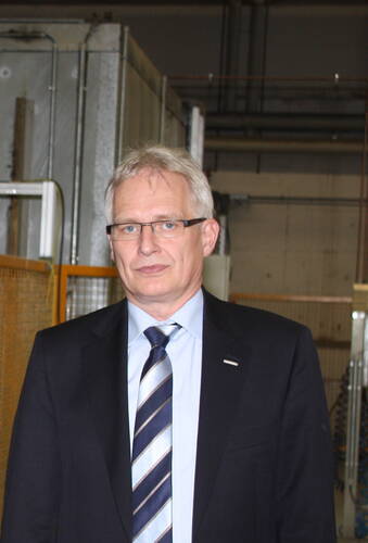 Älterer Mann im Anzug mit gestreifter Krawatte und Brille steht in einer Industriehalle vor technischen Anlagen