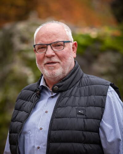 Älterer Mann mit Brille, grauem Bart und gesteppter Weste blickt freundlich in die Kamera vor unscharfem, herbstlichem Hintergrund