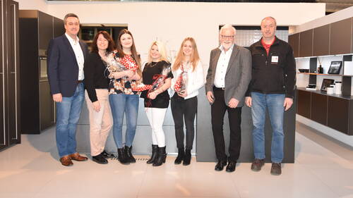 Sieben Personen stehen in einer modernen Küchenausstellung nebeneinander und lächeln in die Kamera, drei junge Frauen in der Mitte halten verpackte Geschenkpräsente in den Händen.