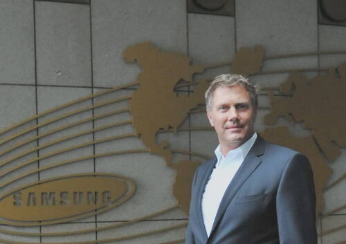 Mittelalter Mann im Anzug steht vor einer Wand mit goldener Weltkarten-Grafik und großem Samsung-Logo