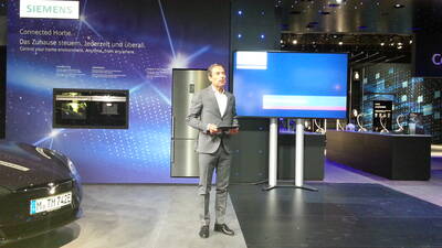 Geschäftsmann in grauem Anzug präsentiert auf einer Siemens-Messebühne ein vernetztes Zuhause mit großem Display und Smart-Home-Geräten im Hintergrund