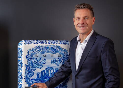 Mann im dunklen Anzug lächelt in moderner Umgebung und lehnt mit der Hand an einem kunstvoll blau-weiß verzierten SMEG-Kühlschrank mit klassischem Motiv