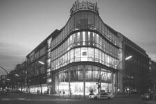 Abendliche Straßenszene mit einem beleuchteten, mehrstöckigen Kaufhaus mit abgerundeter Glasfassade an einer Straßenecke, vor dem Autos und Passanten vorbeiziehen