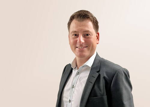 Lächelnder Mann im grauen Anzug und weißem Hemd vor einfarbig hellem Hintergrund in professioneller Porträtpose