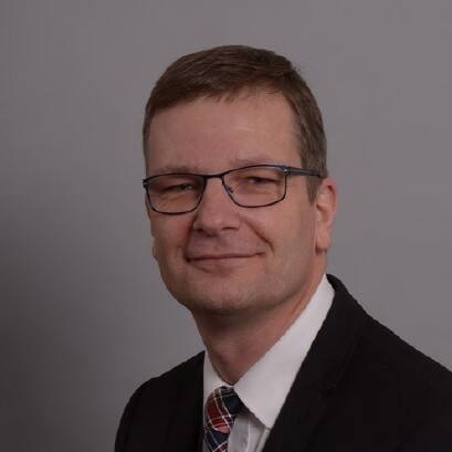 Mittelalterlicher Mann mit Brille und kurzem braunem Haar, trägt dunklen Anzug, weißes Hemd und karierte Krawatte, lächelt leicht vor einfarbig grauem Hintergrund