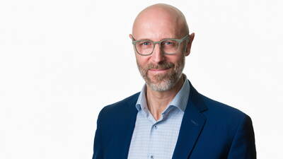 Glatzköpfiger Mann mit Brille und grauem Bart lächelt im blauen Anzug vor weißem Hintergrund