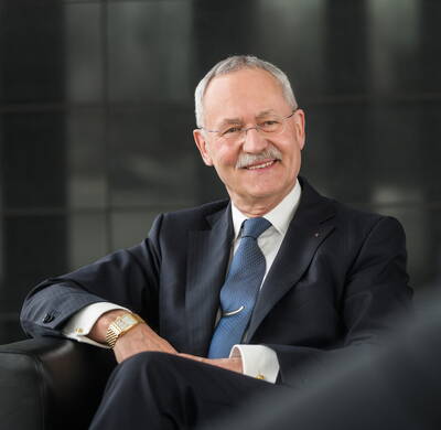 Älterer Mann im dunklen Anzug sitzt entspannt auf einem Ledersessel, lächelt freundlich und blickt leicht zur Seite.