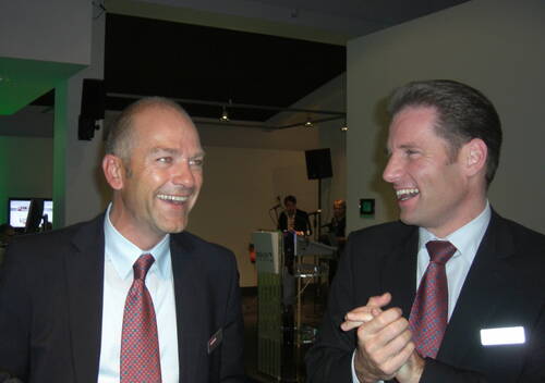 Zwei geschäftlich gekleidete Männer im Anzug lachen und unterhalten sich angeregt bei einer Firmenveranstaltung in einem modern beleuchteten Innenraum.