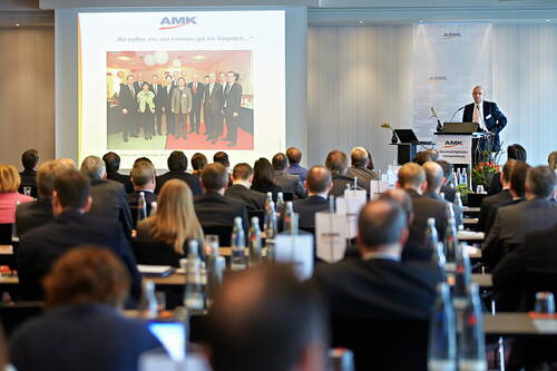 Redner präsentiert vor voll besetztem Konferenzsaal eine Folie mit Gruppenfoto auf großer Leinwand.