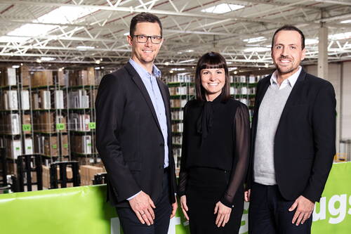 Drei lächelnde Führungskräfte in Businesskleidung stehen vor Hochregalen in einem modernen Logistiklager