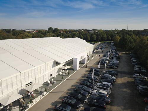 Großes weißes Eventzelt mit überdachtem Eingangsbereich und dicht belegtem Parkplatz, umgeben von Bäumen und blauem Himmel in einer ländlichen Umgebung