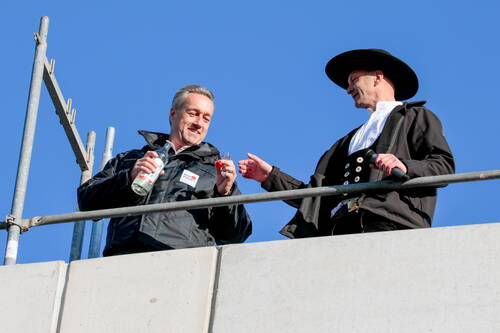 Zwei Männer stoßen auf einem Rohbaugerüst mit Schnapsgläsern an, einer trägt traditionelle Zimmermannskleidung mit Hut, der andere eine Arbeitsjacke mit Flasche in der Hand.