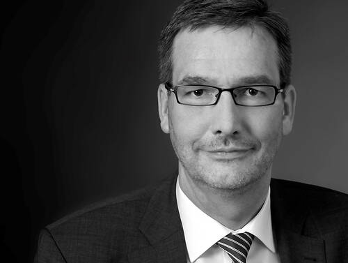 Seriöser Mann mittleren Alters mit Brille im Anzug und gestreifter Krawatte blickt ruhig in die Kamera vor dunklem Hintergrund.