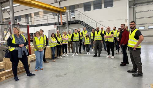 Gruppe von Mitarbeitenden in gelben Warnwesten hört in einer Lagerhalle einer Kollegin zu, die vor ihnen spricht