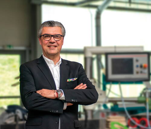 Selbstbewusster Geschäftsmann im Anzug steht mit verschränkten Armen in einer modernen Industriehalle vor einer unscharfen Maschinensteuerung