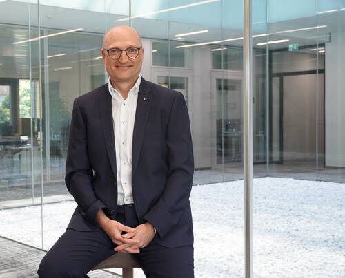 Mittelalterlicher Mann im Anzug sitzt lächelnd in einem modernen Büro mit Glaswänden und hellem, steinbedecktem Innenhof im Hintergrund