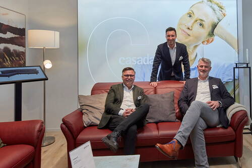Drei Geschäftsleute im Anzug präsentieren entspannt eine rote Ledersofa-Garnitur in modernem Showroom mit großem Marken-Display im Hintergrund.