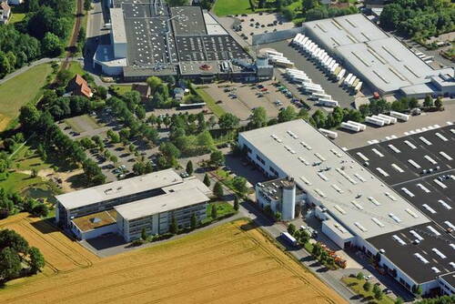 Großes Industrie- und Logistikgelände mit mehreren Hallen, Parkplätzen und Lkw-Ladezonen am Rand von Feldern und Grünflächen aus der Luftperspektive betrachtet.