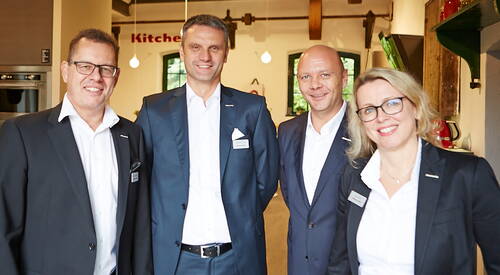 Vier lächelnde Geschäftsleute in Anzügen posieren nebeneinander in einem modernen, hellen Büro- oder Ausstellungsraum.