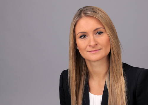 Junge Frau mit langen blonden Haaren und schwarzem Blazer blickt freundlich in die Kamera vor neutral grauem Hintergrund
