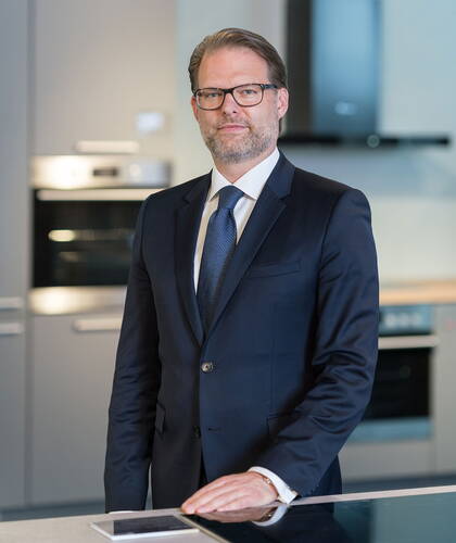 Geschäftsmann im Anzug steht in einer modernen Küche und stützt eine Hand auf die Arbeitsplatte