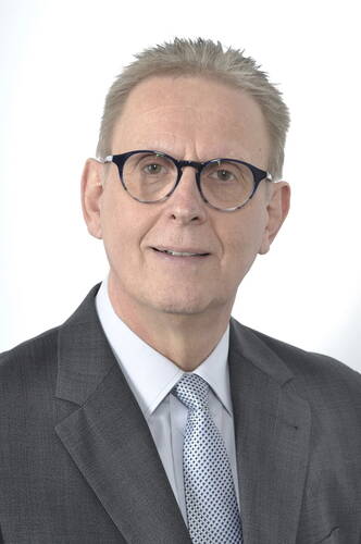 Älterer Mann mit kurzer, hellgrauer Frisur, dunkler Brille und grauem Anzug mit gemusterter Krawatte vor weißem Hintergrund