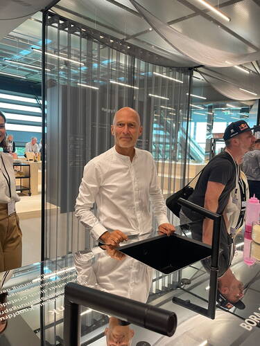 Mann in weißem Hemd steht lächelnd an einem modernen Küchenstand mit schwarzer Spüle in einer hellen, futuristischen Ausstellungshalle.