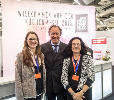 Drei lächelnde Messemitarbeitende mit Namensschildern posieren gemeinsam vor dem Willkommensbanner der Küchenmesse 2015 am Informationsstand.