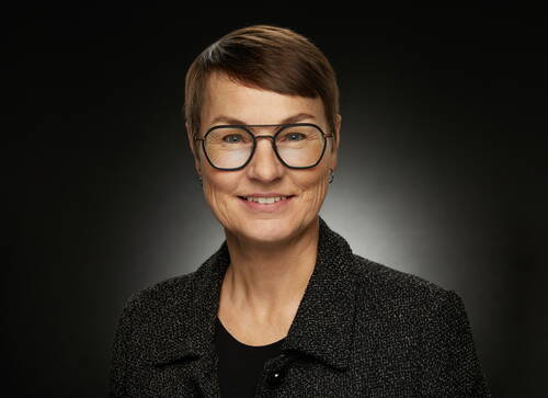 Lächelnde Frau mit Kurzhaarfrisur und großer Brille in dunkler Jacke vor neutral dunklem Hintergrund
