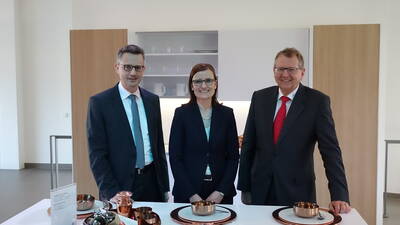 Drei geschäftlich gekleidete Personen stehen lächelnd in einer modernen Ausstellungsküche hinter einem Tisch mit Kupfergeschirr und Gedecken.
