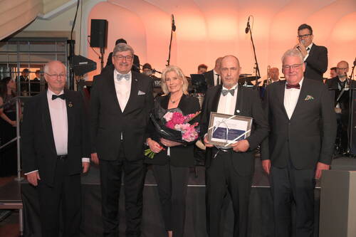 Fünf elegant gekleidete Personen posieren auf einer Bühne vor einer Gala-Band, während eine Frau einen Blumenstrauß und ein Mann eine gerahmte Urkunde als Auszeichnungen hält.