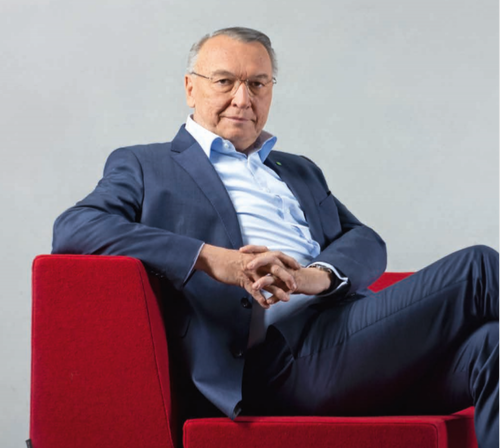 Älterer Mann im Business-Anzug sitzt entspannt in rotem Sessel und schaut direkt in die Kamera