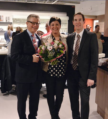 Drei festlich gekleidete Personen lächeln in einem modernen Innenraum in die Kamera, während die Frau in der Mitte einen großen Blumenstrauß als Geschenk hält.