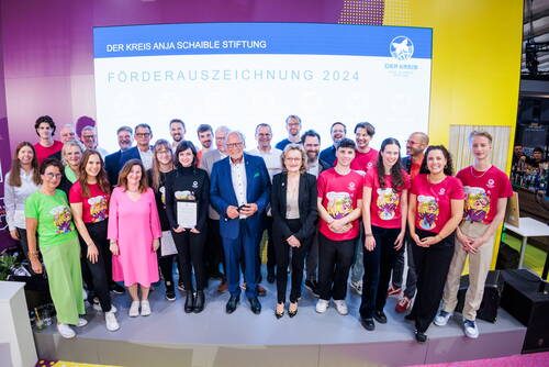 Gruppenfoto der Preisträgerinnen und Preisträger der Förderauszeichnung 2024 der DER KREIS Anja Schaible Stiftung auf einer Bühne, umgeben von Vertretern der Stiftung und jungen Menschen in bunten T-Shirts.