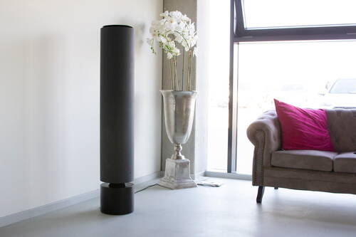 Modernes Wohnzimmer mit schlankem schwarzen Standlautsprecher, silberner Vase mit weißen Orchideen und beigem Chesterfield-Sofa mit auffälligem pinkem Kissen vor einer großen Fensterfront