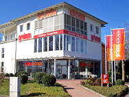 Moderne Küchenausstellung in einem hellen Eckgebäude mit großen Glasfronten, roter Beschriftung „Küchen“ und bunten Fahnen an einem sonnigen Tag