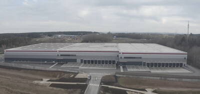 Großes, modernes Logistikzentrum mit zahlreichen Laderampen vor bewaldeter und leicht hügeliger Umgebung unter bedecktem Himmel