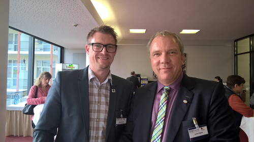 Zwei Geschäftsleute im Anzug lächeln in die Kamera, während sie bei einer Konferenz im Foyer mit Stehtischen und weiteren Teilnehmenden im Hintergrund stehen.