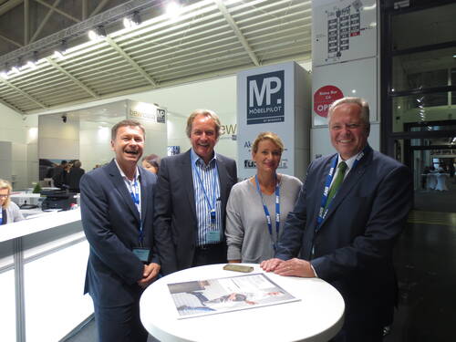 Vier lächelnde Messebesucher stehen an einem runden Stehtisch vor dem Messestand von „Möbelpilot“ in einer hell beleuchteten Ausstellungshalle.
