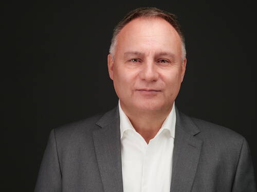 Reifer Mann im grauen Anzug und weißem Hemd blickt mit neutralem Gesichtsausdruck vor dunklem Hintergrund direkt in die Kamera